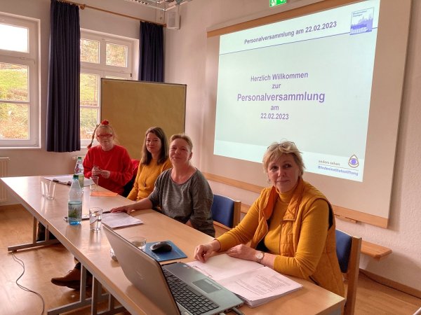 Vier Frauen sitzen an einem Tisch und schauen in die Kamera. Vor den Frauen sind ein Laptop, Dokumente und Wasserflaschen platziert. Hinter ihnen wird eine Power-Point-Präsentation mit dem Titel Herzlich Willkommen zur Personalversammlung am 22.02.23 projiziert.