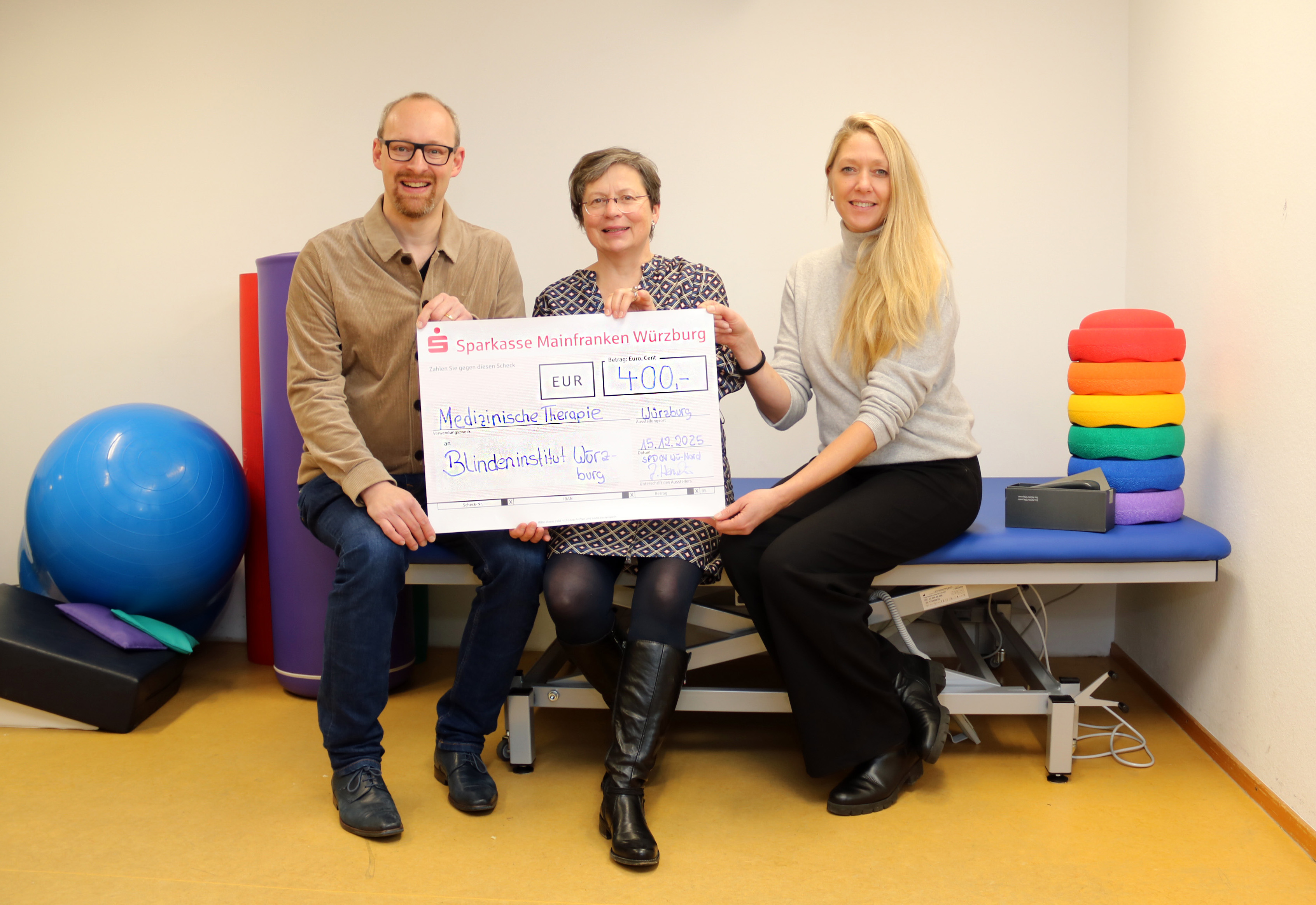 Ein Mann und zwei Frauen sitzen auf einer Therapieliege und halten einen Spendenscheck in der Hand