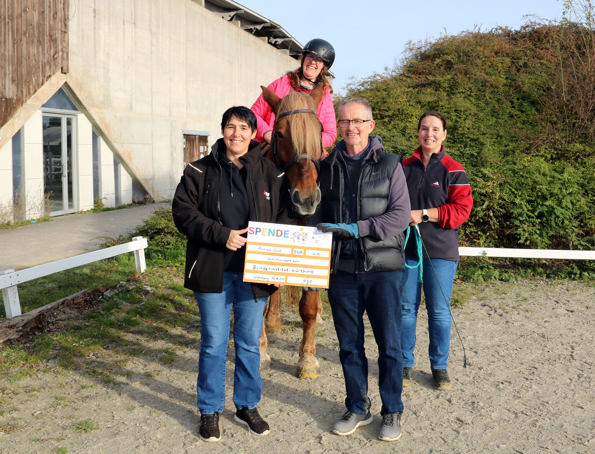 Zwei Frauen und ein Mann stehen neben einem Pferd, eine junge Frau sitzt auf dem Pferd. Eine Frau und der Mann halten einen Spendenscheck in der Hand
