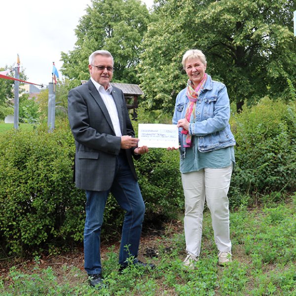 Zwei erwachsene Personen stehen auf einer Wiese mit Büschen im Hintergrund.Links im Bild steht Reinhold Stabel und hält mit Annette Peters, die rechts neben ihm steht, einen Spendenscheck in der Hand.