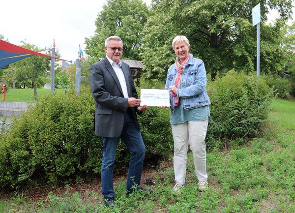 Zwei erwachsene Personen stehen auf einer Wiese mit Büschen im Hintergrund.Links im Bild steht Reinhold Stabel und hält mit Annette Peters, die rechts neben ihm steht, einen Spendenscheck in der Hand.