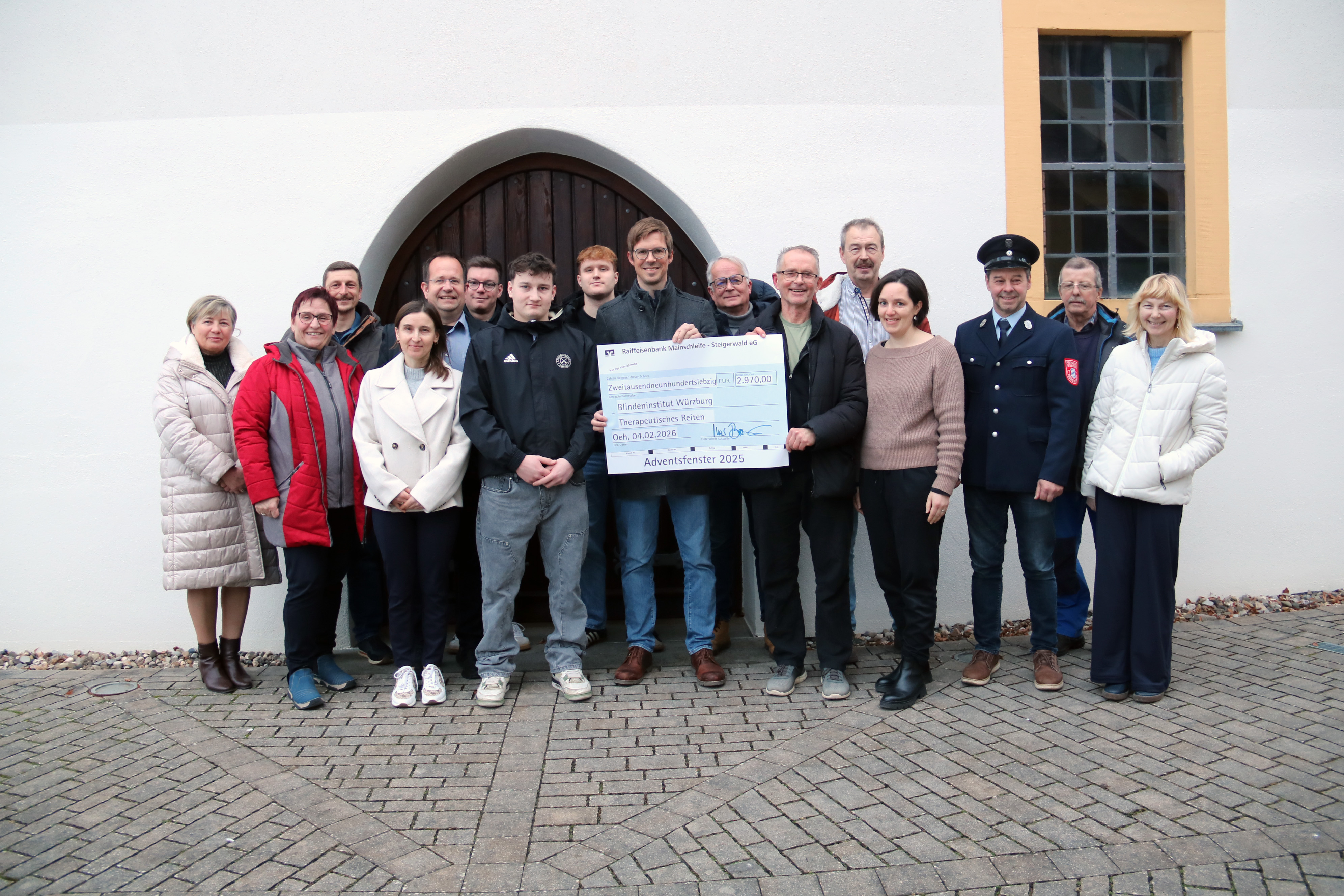 Sechzehn Personen stehen als Gruppe vor einer Kirche, Zwei davon halten einen Spendenscheck in der Mitte.