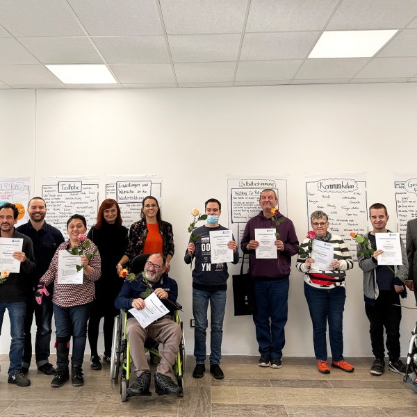 Eine Grupper mit 13 Männer und Frauen mit und ohne Behinderung steht im Halbkreis vor einer Wand und acht Personen halten ein Zertifikat in den Händen.