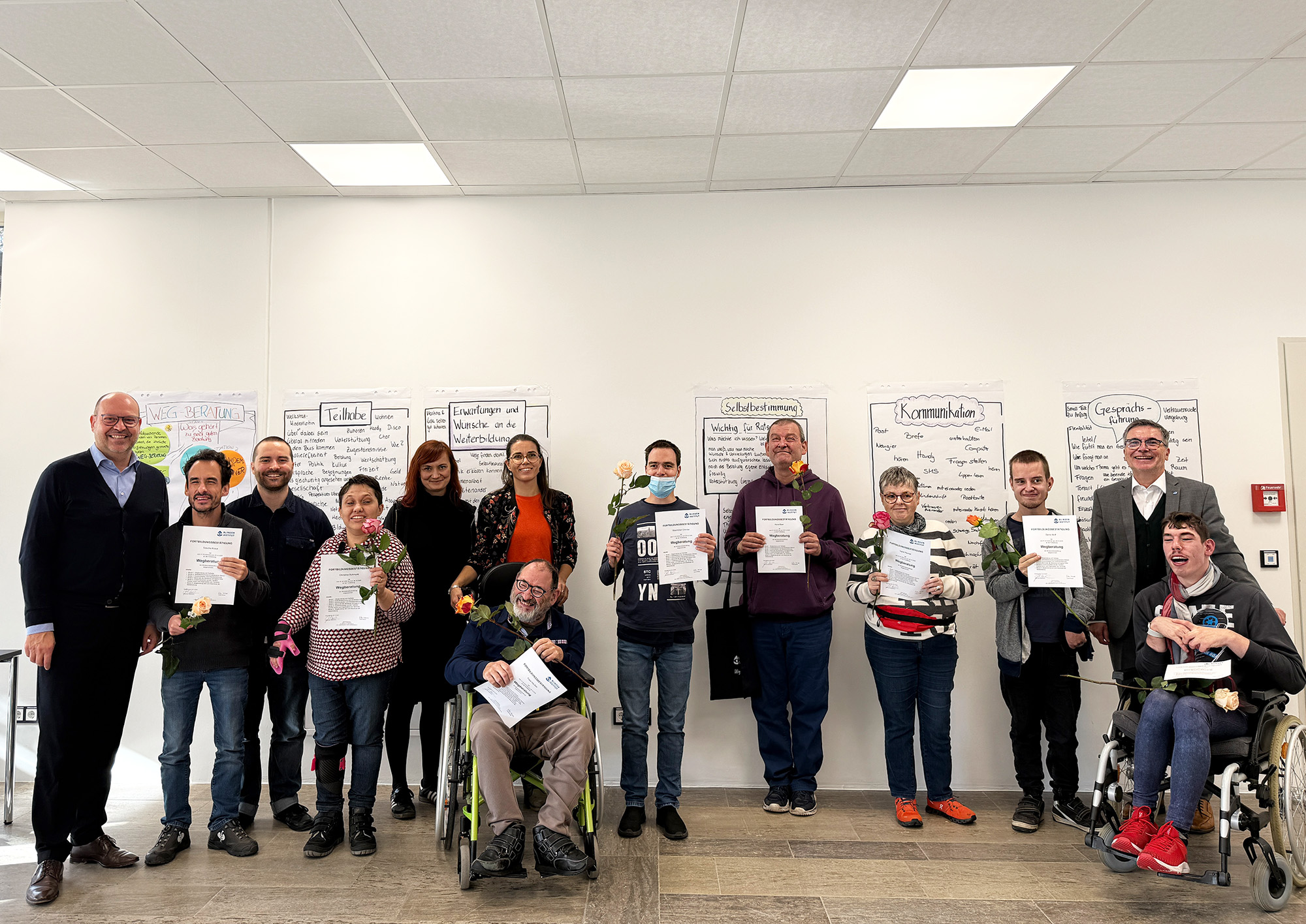 Eine Grupper mit 13 Männer und Frauen mit und ohne Behinderung steht im Halbkreis vor einer Wand und acht Personen halten ein Zertifikat in den Händen.