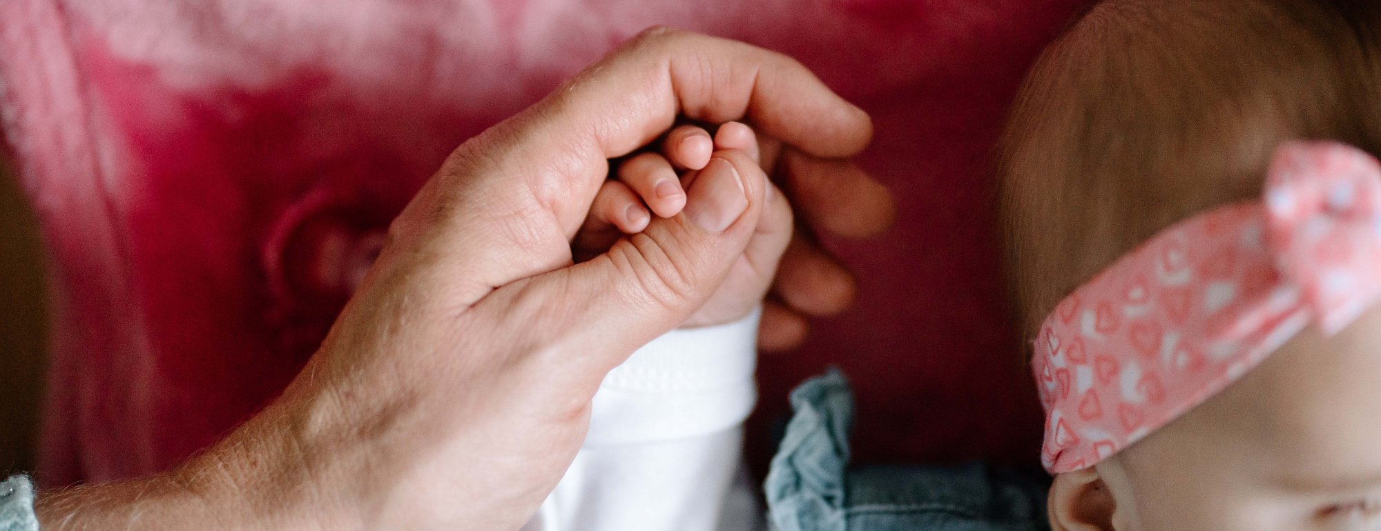 Auf dem Bild ist die Hand einer Frau zu sehen. Sie hält die Hand eines Babys, das auf einer rosanen Kuscheldecke liegt.