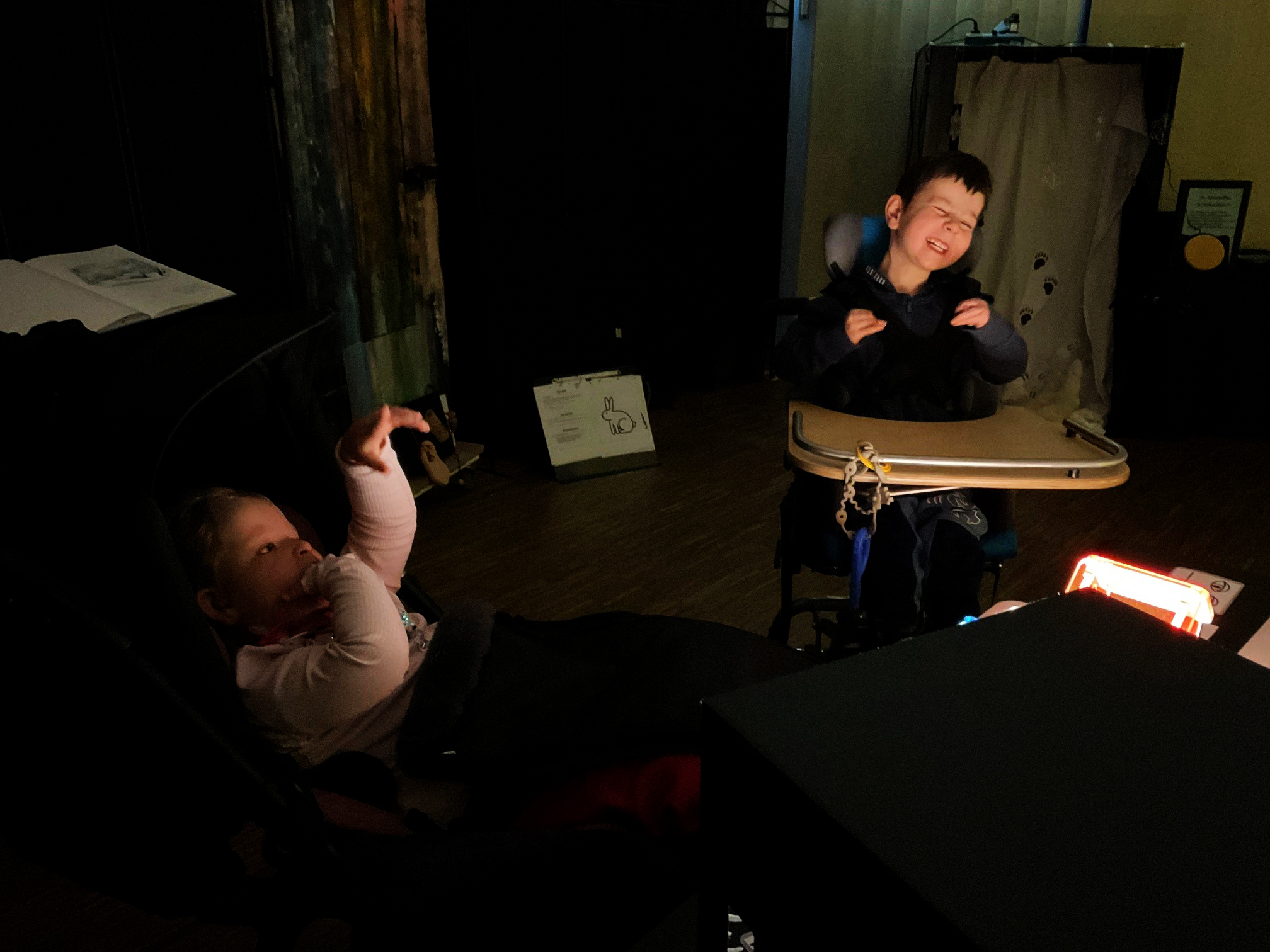 Zwei Kinder sitzen in einem abgedunkelten Raum, eines in einem Liegesitz und eines an einem Tisch mit Ablagefläche. Vor ihnen steht eine leuchtende Lichtquelle. Im Hintergrund sind Wandvorhänge, ein offenes Buch und verschiedene Materialien für ein Sinnes- oder Sehangebot zu sehen.