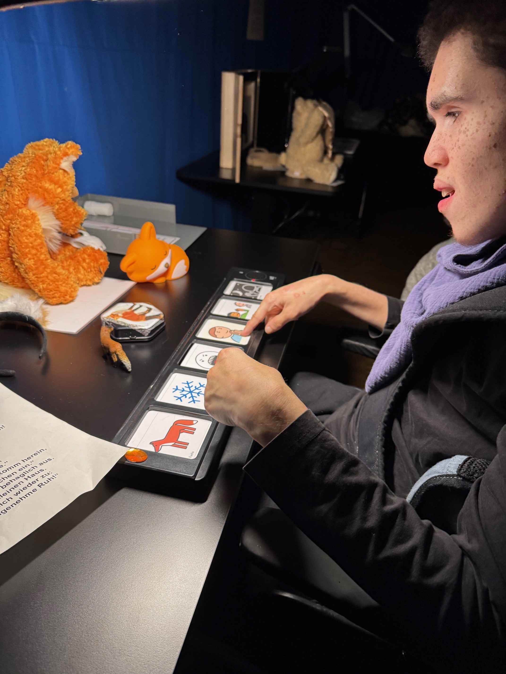 Hände ertasten eine Reihe taktiler Symbolkarten auf einem Tisch, umgeben von verschiedenen fühlbaren Tierfiguren und Materialien.