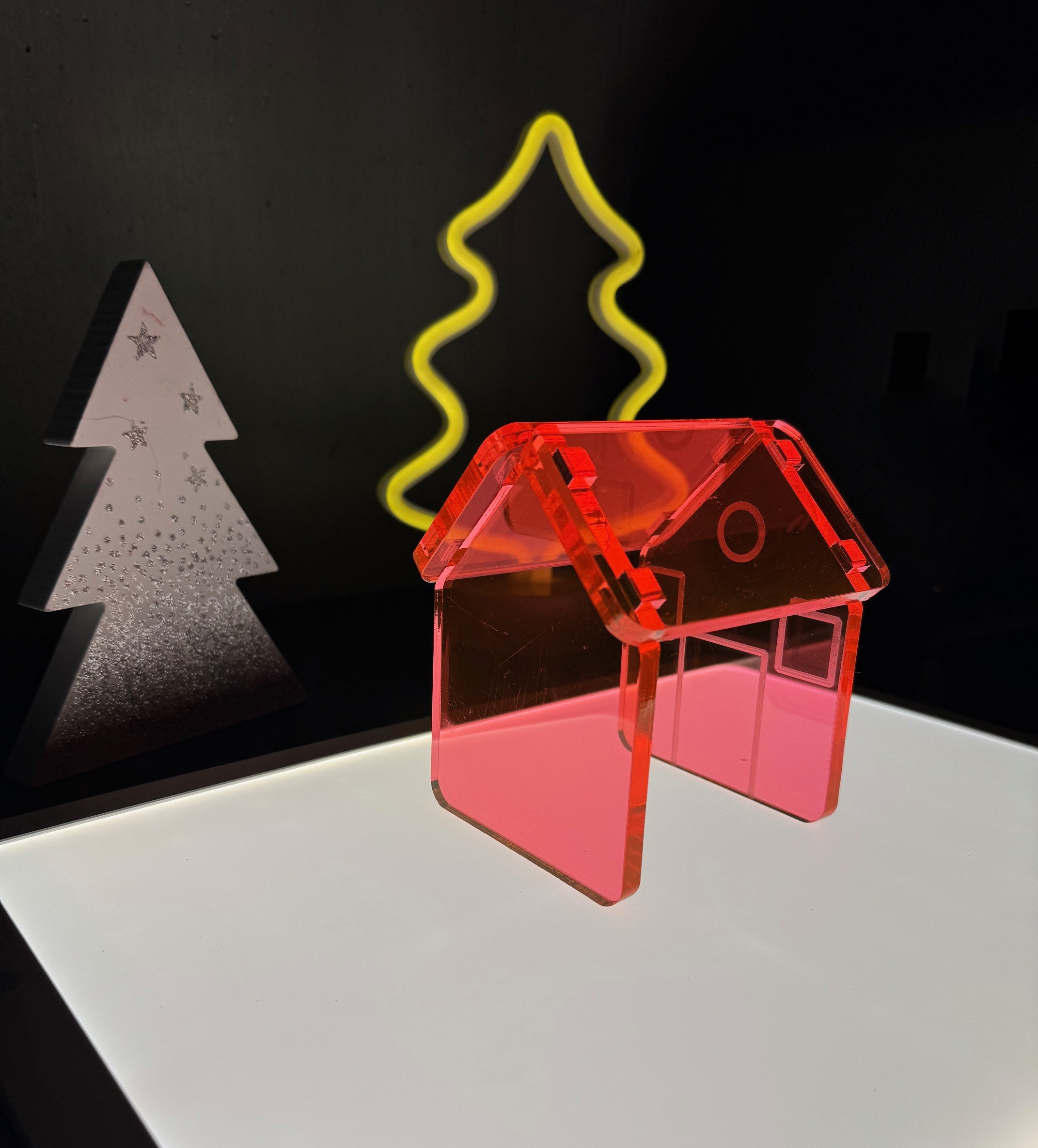 Auf einem hell beleuchteten Leuchttisch steht ein kleines transparentes rotes Miniaturhaus aus Kunststoff. Dahinter sind zwei stilisierte Tannenbäume zu sehen – einer aus mattem Material, der andere als gelb leuchtende Kontur. Die Szene ist dunkel umgeben, sodass die Objekte klar hervortreten.