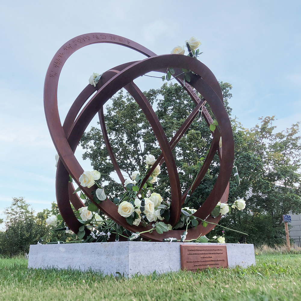Rostfarbene Metallskulptur aus verschlungenen Ringen auf einem Sockel im Freien, geschmückt mit weißen Rosen. Dahinter stehen Bäume und Wiese.

