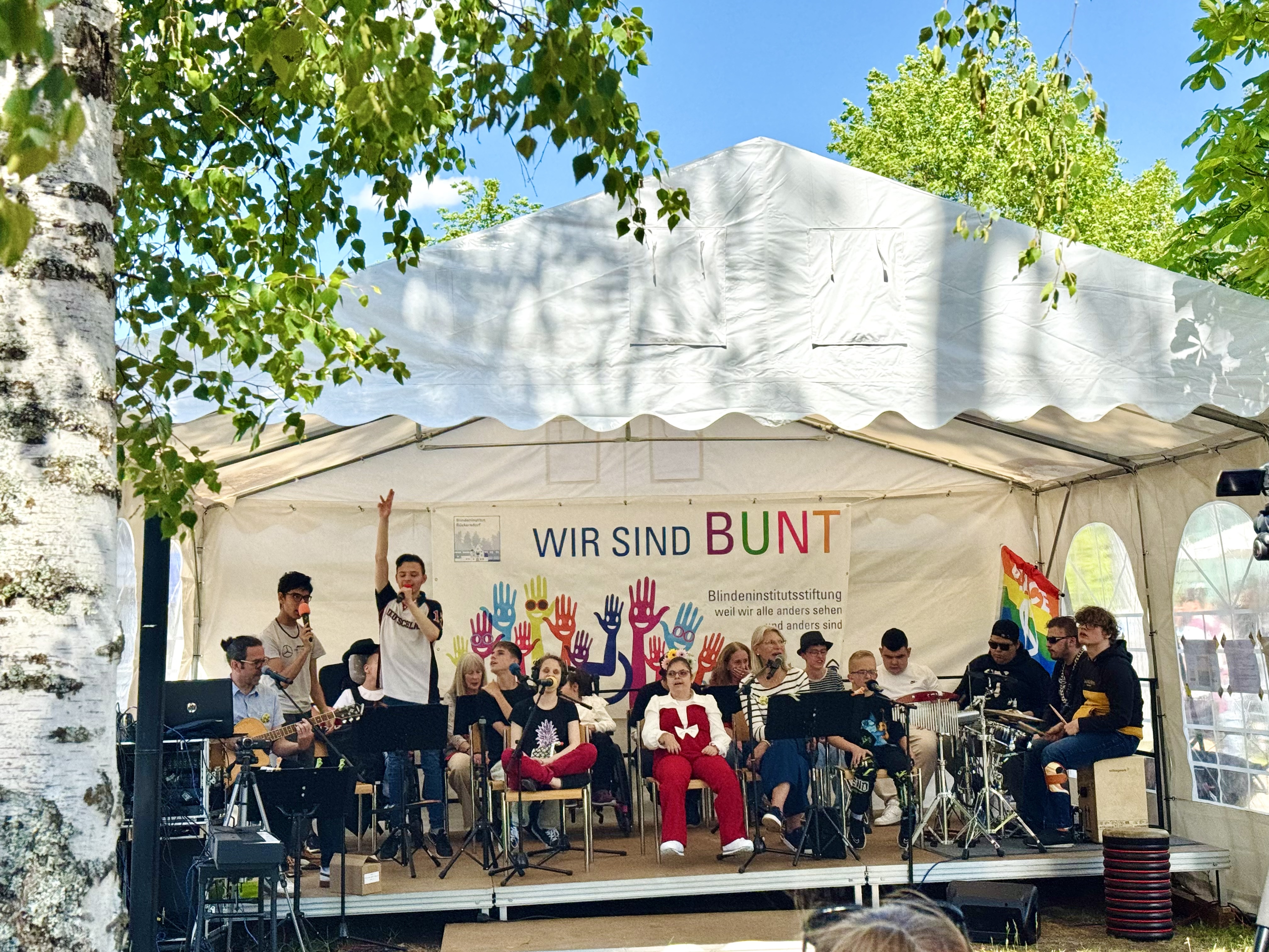 Die Schülerband „Studio D“ tritt auf einer Bühne in einem weißen Festzelt beim Sommerfest auf. Die Musikerinnen und Musiker spielen verschiedene Instrumente und singen gemeinsam vor Publikum. Hinter der Band hängt ein Banner mit der Aufschrift „WIR SIND BUNT“ und bunten Handabdrücken. Rechts neben der Bühne ist eine Regenbogenfahne zu sehen. Das Zelt steht unter grünen Bäumen an einem sonnigen Tag.