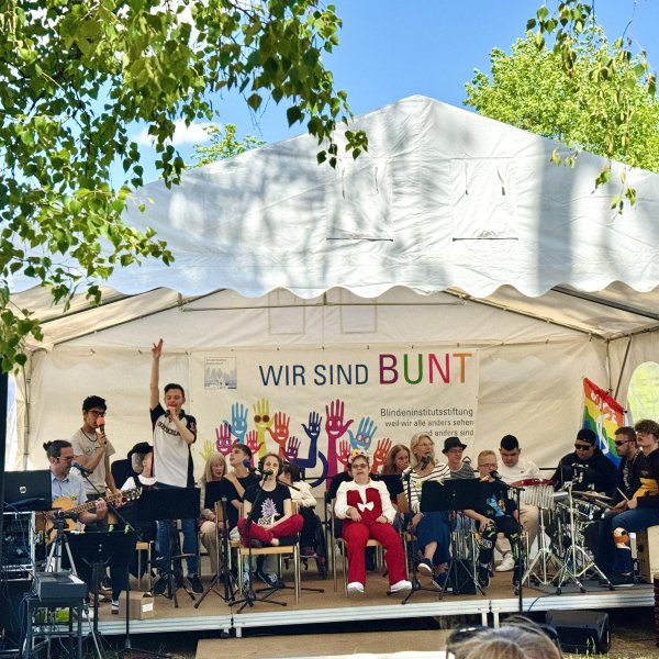 Die Schülerband „Studio D“ tritt auf einer Bühne in einem weißen Festzelt beim Sommerfest auf. Die Musikerinnen und Musiker spielen verschiedene Instrumente und singen gemeinsam vor Publikum. Hinter der Band hängt ein Banner mit der Aufschrift „WIR SIND BUNT“ und bunten Handabdrücken. Rechts neben der Bühne ist eine Regenbogenfahne zu sehen. Das Zelt steht unter grünen Bäumen an einem sonnigen Tag.