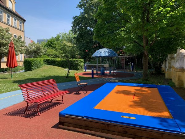 Das Bild zeigt einen Ausschnitt des Spielplatzes am Blindeninstitut. Im Vordergrund sieht man das Trampolin und daneben eine rote Bank. Im Hintergrund sieht man das Gebäude, einen roten Schirm und ein Karusell.