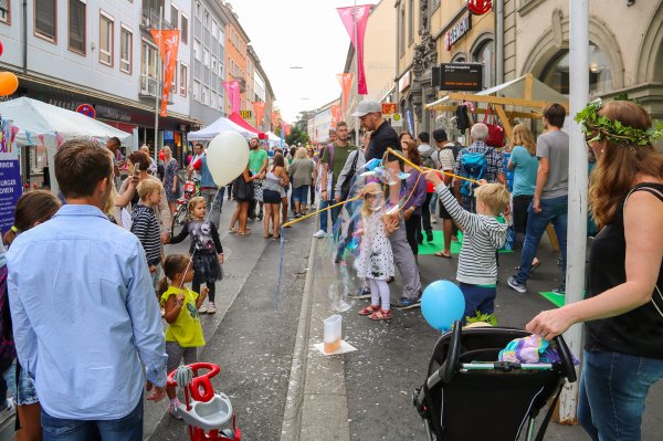 Kinder machen bei einem Straßenfest große Seifenblasen, während im Hintergrund viele Menschen zwischen Ständen und Fahnen durch eine Fußgängerzone gehen.