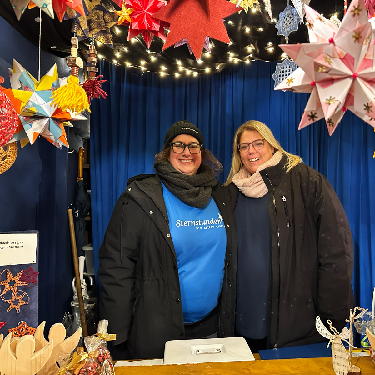 Sternstunden-Stand mit zwei Helferinnen, dekoriert mit bunten Sternen und Lichterketten, vor einem Tisch mit Weihnachtsartikeln.“

Bild 2
Bildbeschreibung:
„Drei Helferinnen am Sternstunden-Stand auf dem Nürnberger Christkindlesmarkt. Über ihnen hängen zahlreiche bunte Sterne und Lichterketten, auf dem Tisch liegen Spendenboxen, ein Korb und handgefertigte Weihnachtsdekorationen.“
Alt-Text:
„Sternstunden-Stand mit drei Helferinnen, umgeben von bunten Sternen und Lichterketten, vor einem Tisch mit Spendenbox und Weihnachtsartikeln.