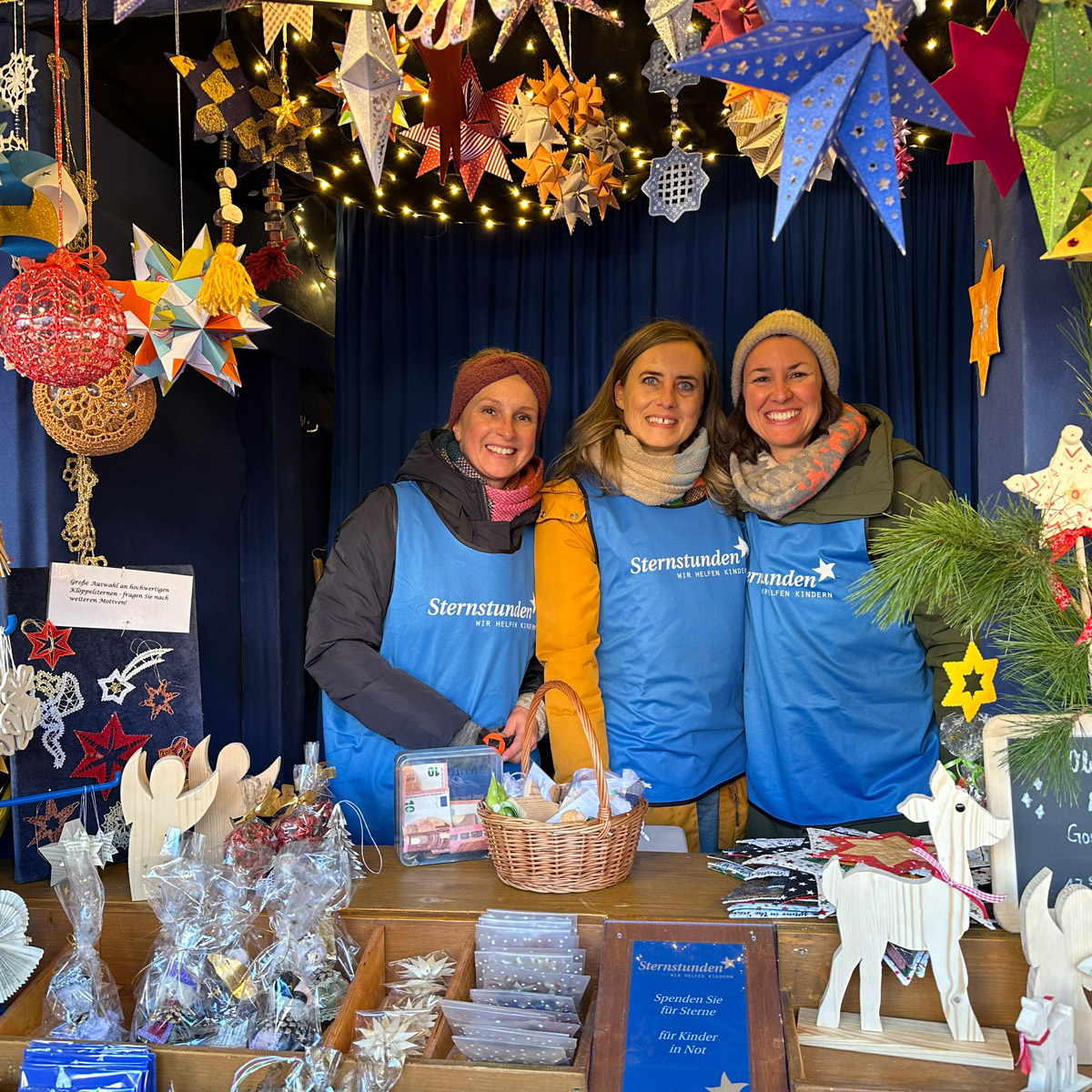 Drei Helferinnen am Sternstunden-Stand auf dem Nürnberger Christkindlesmarkt. Über ihnen hängen zahlreiche bunte Sterne und Lichterketten, auf dem Tisch liegen Spendenboxen, ein Korb und handgefertigte Weihnachtsdekorationen.“