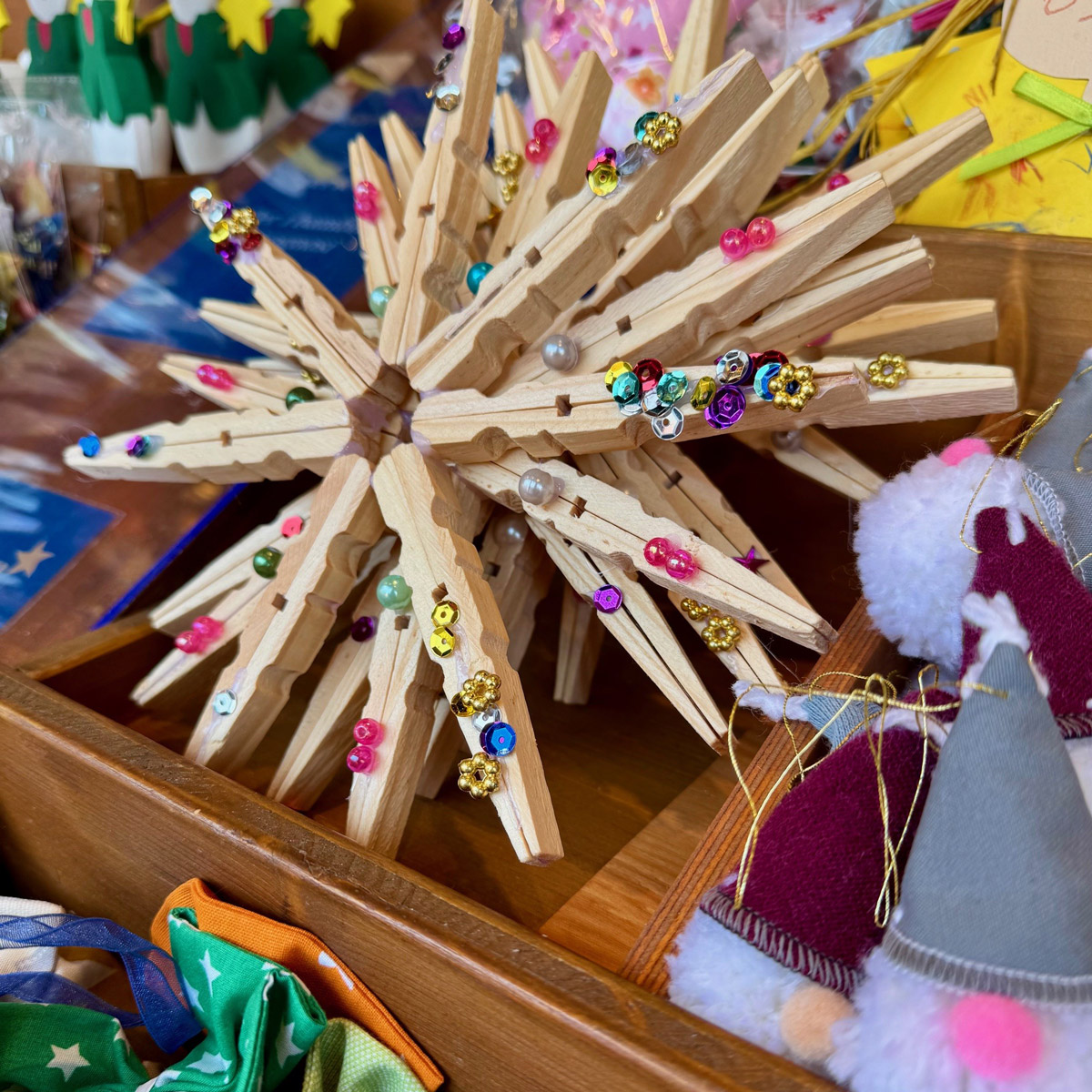 Handgefertigter Stern aus Holz-Wäscheklammern, verziert mit bunten Perlen und Glitzer, auf einem Marktstand mit weiteren Weihnachtsdekorationen.