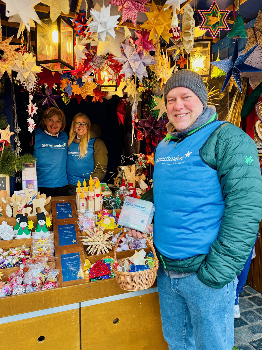 Sternstunden-Stand auf dem Nürnberger Christkindlesmarkt mit bunten, handgefertigten Sternen und weihnachtlichen Dekorationen. Auf dem Tisch liegen Sterne, Holzfiguren und kleine Geschenkartikel, dahinter Helfer*innen in blauen Sternstunden-Schürzen.