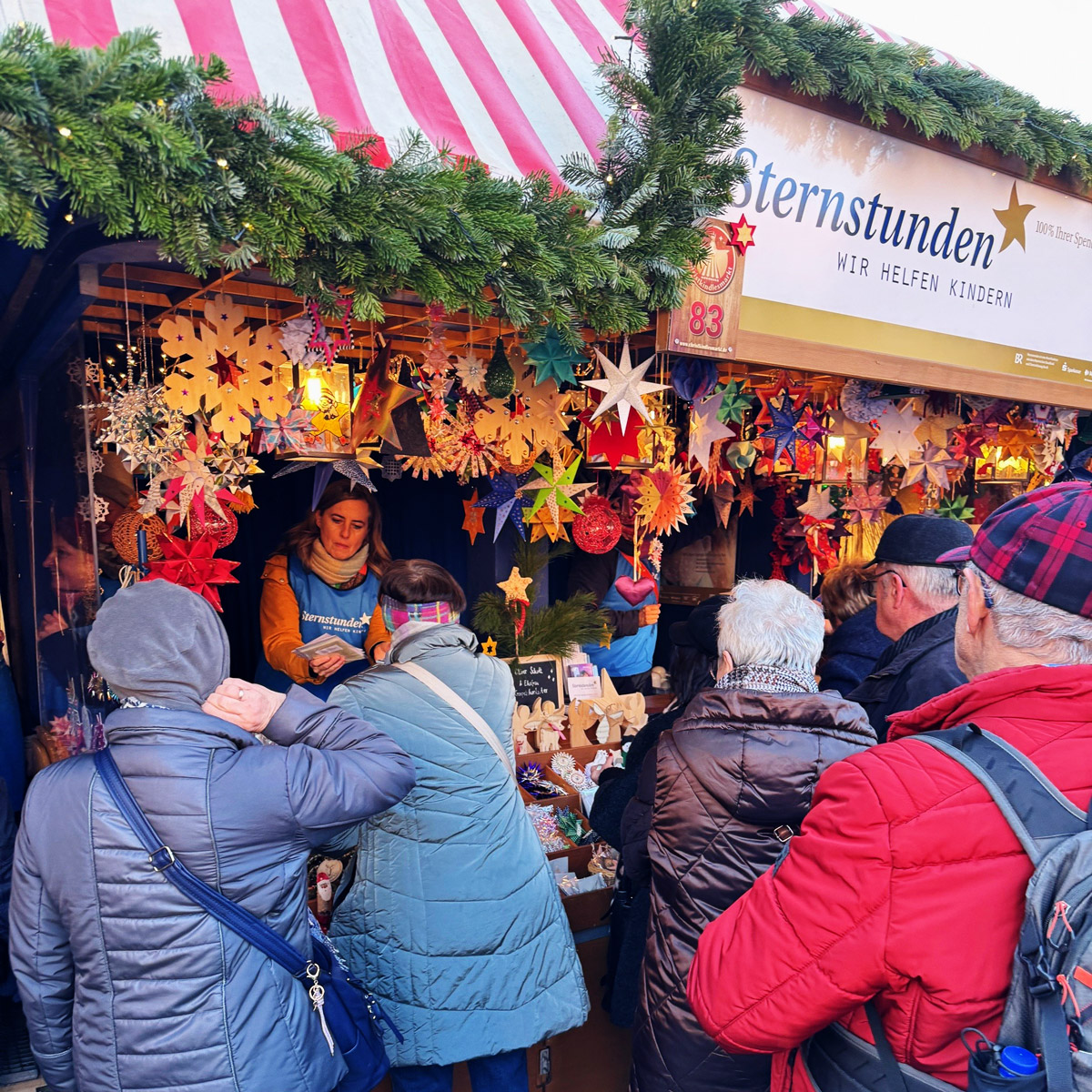 Sternstunden-Stand auf dem Christkindlesmarkt mit bunten Sternen und Weihnachtsdekorationen, mehrere Besucher davor.