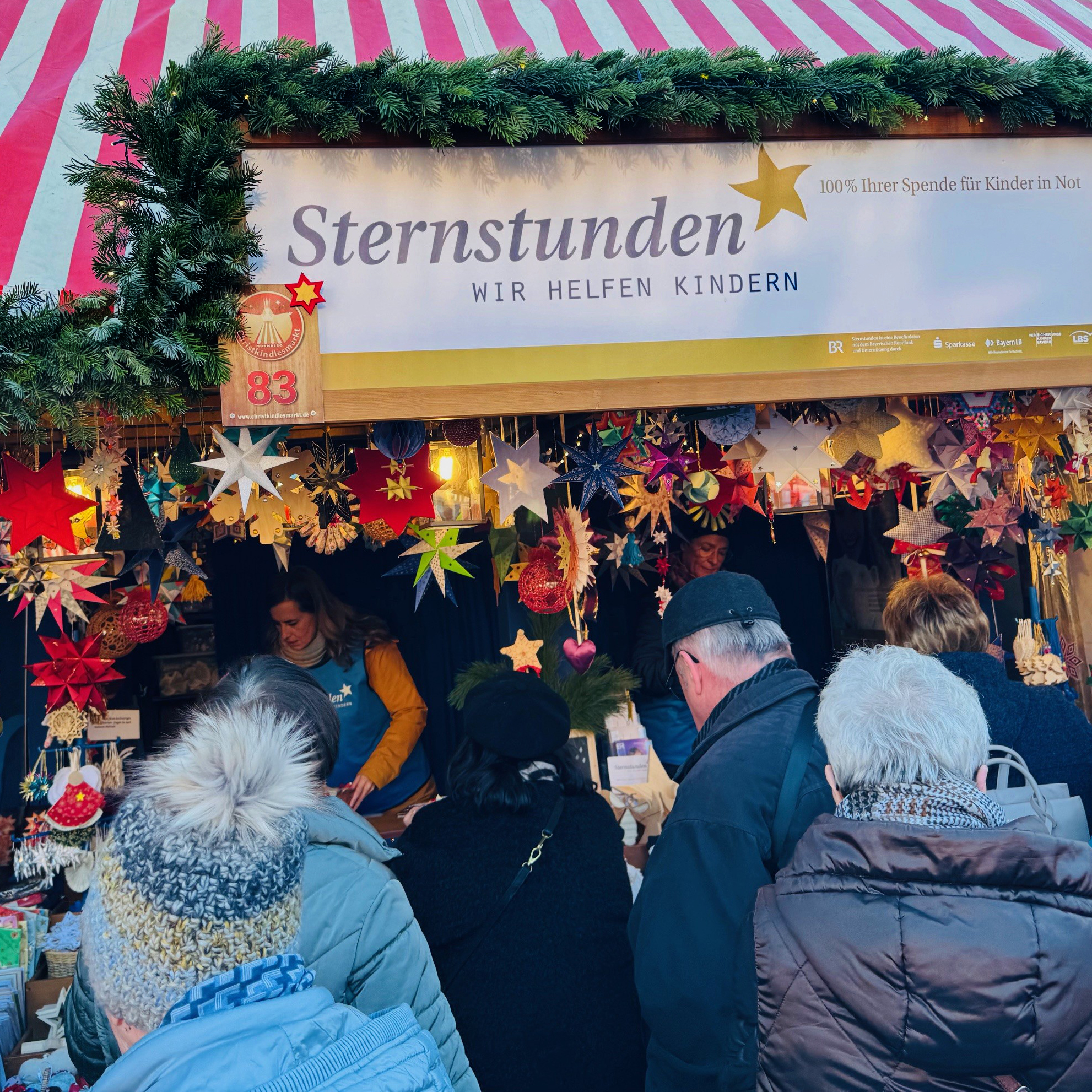Sternstunden-Stand auf dem Nürnberger Christkindlesmarkt mit vielen bunten Sternen und Weihnachtsdekorationen. Mehrere Besucher stehen vor dem Stand, über dem ein Schild mit der Aufschrift ‚Sternstunden – Wir helfen Kindern‘ hängt.
