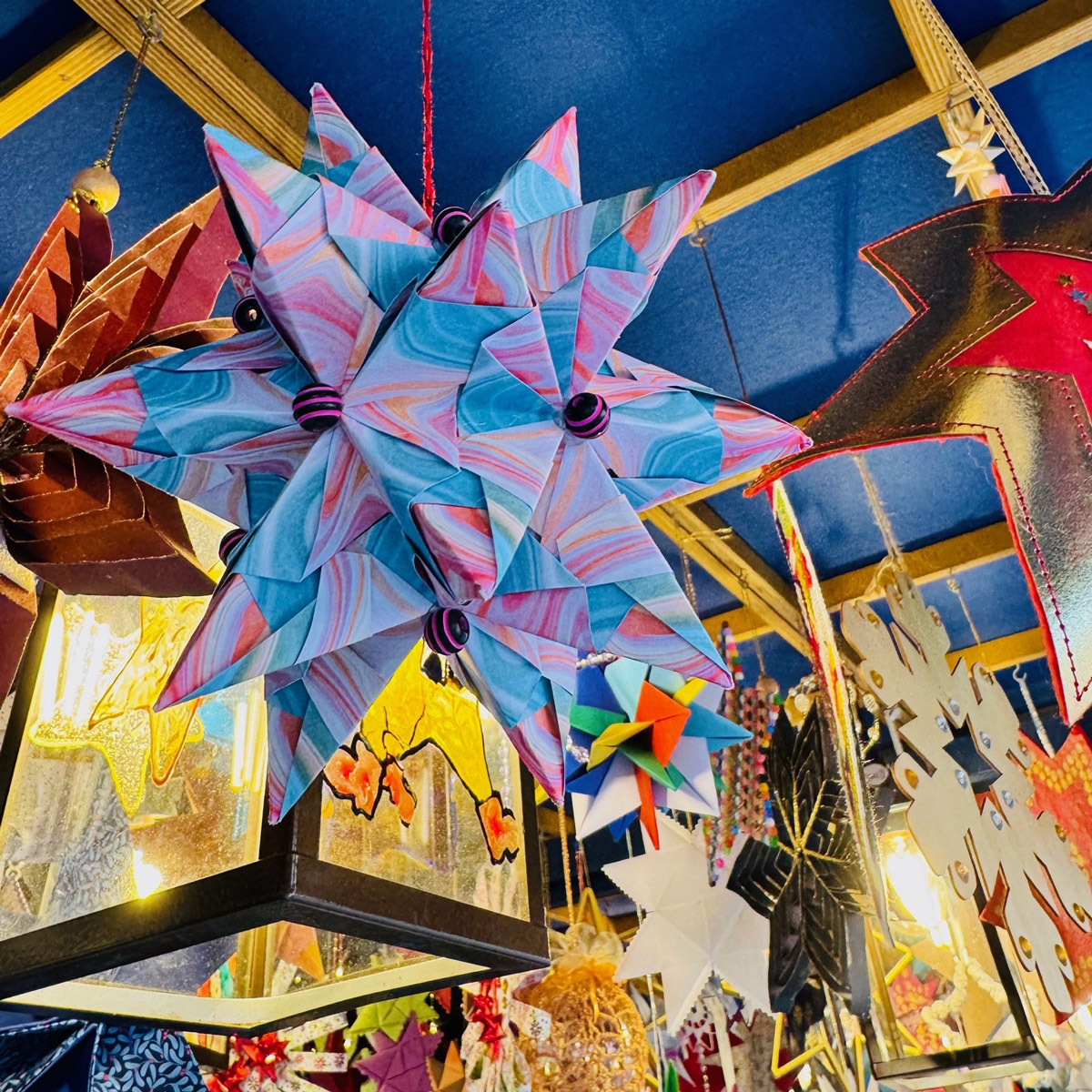 Großer, bunter Papierstern mit rosa und blauen Mustern, umgeben von weiteren Sternen und Lichterketten am Marktstand.