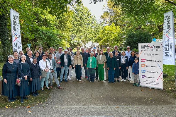 Eine große Gruppe von Erwachsenen und einem Kind steht für ein gemeinsames Foto auf einem Weg in einem grünen Park. Rechts neben der Gruppe stehen ein Roll-up und zwei Fahnen des Würzburger Stiftungsnetzwerks.