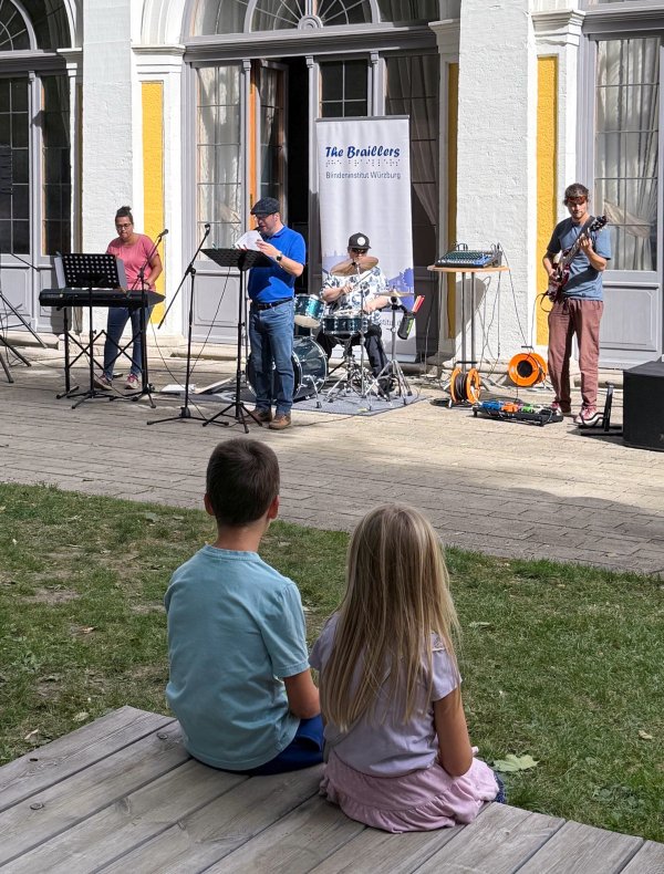 Zwei Kinder sitzen mit dem Rücken zur Kamera auf einer Holzbank und schauen einer vierköpfigen Band bei einem Auftritt im Freien zu. Hinter der Band steht ein Banner mit der Aufschrift „The Braillers – Blindeninstitut Würzburg“.