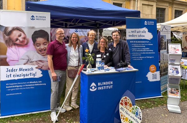 Fünf Erwachsene stehen lächelnd an einem Informationsstand des Blindeninstituts unter einem blauen Pavillon. Der Stand ist mit Roll-ups, Infomaterialien und einem Stehtisch ausgestattet.