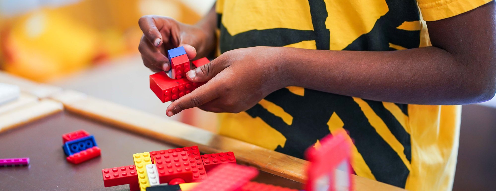 Ein Kind hält bunte Legosteine in den Händen. Vor dem Kind befindet sich ein aus Legosteinen gebautes Objekt, sowie weitere auf dem Tisch verteilte Legosteine.