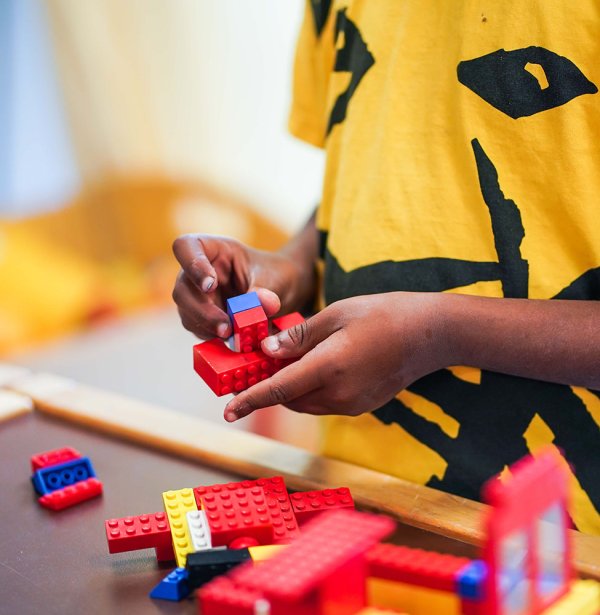 Ein Kind hält bunte Legosteine in den Händen. Vor dem Kind befindet sich ein aus Legosteinen gebautes Objekt, sowie weitere auf dem Tisch verteilte Legosteine.