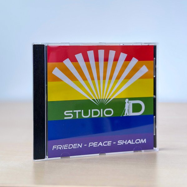 Eine CD-Hülle mit Cover-Motiv: Auf dem Cover steht Studio D - Frieden, Peace, Shalom. Im Hintergrund sind farbige Balken in den Farben des Regenbogens.