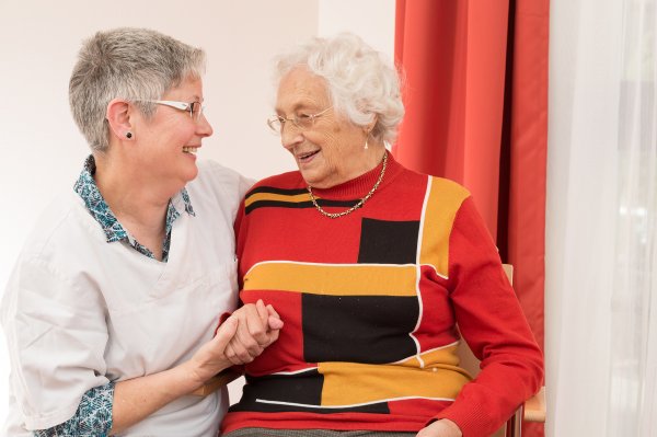 Das Bild zeigt eine Pflegerin und eine Seniorin. Beide sind einander zugewandt und halten sich an den Händen.