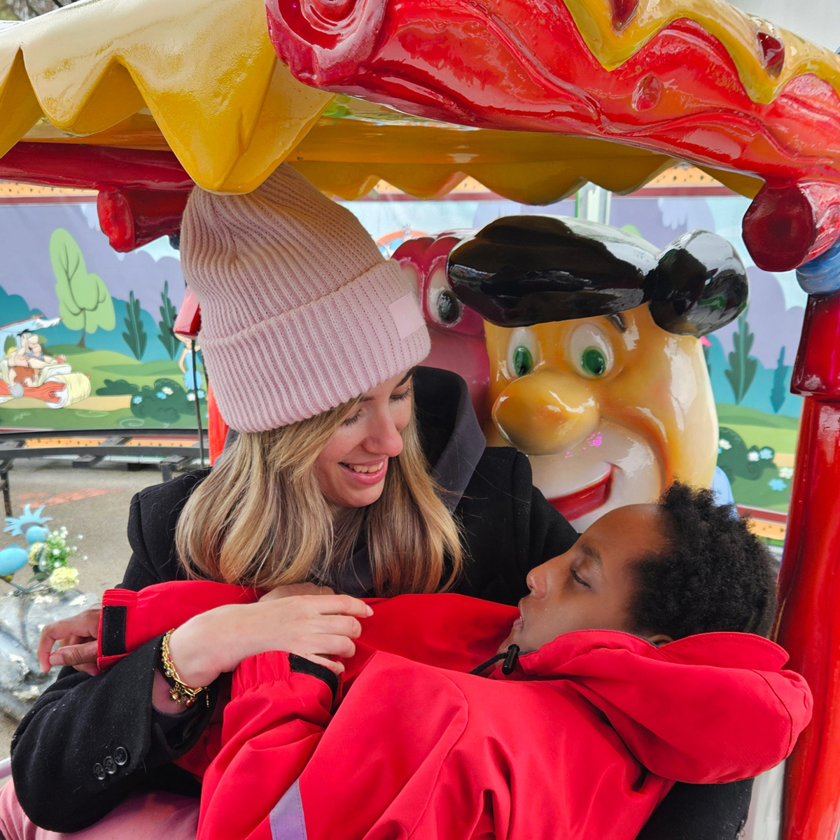 Eine Betreuungsperson sitzt mit einem Kind in einem Fahrgeschäft auf dem Nürnberger Volksfest. Das Kind trägt eine rote Jacke und liegt angegurtet im Schoß der Betreuungsperson. Das bunte Karussell ist mit Figuren und leuchtenden Farben gestaltet.