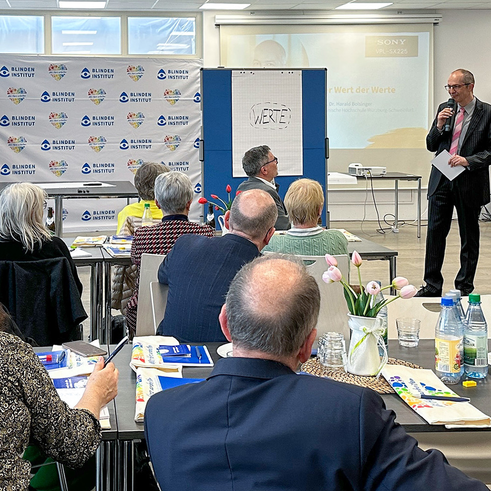 Ein Mann steht vorne im Raum und spricht zu einer Gruppe von Menschen. Die Menschen sitzen an Tischen und hören zu. Auf einer Tafel steht das Wort Werte. Im Hintergrund ist eine Präsentation zu sehen.