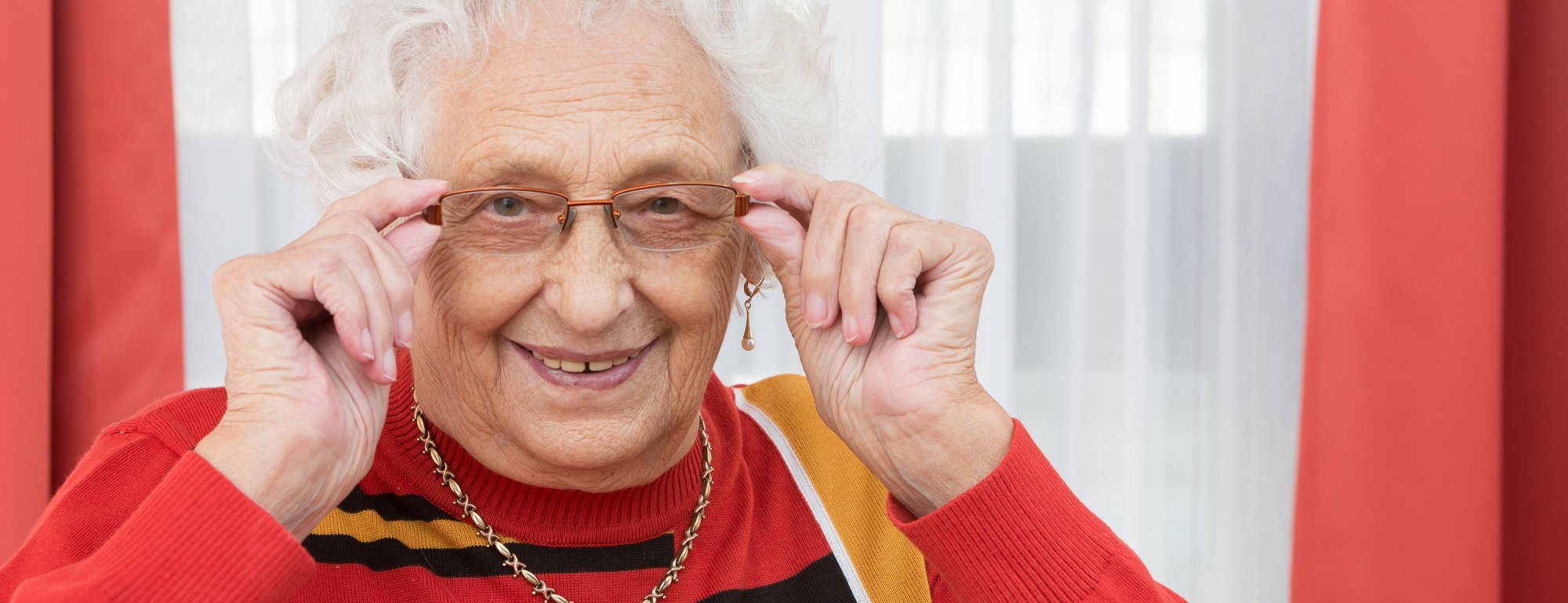 Das Bild zeigt eine weißhaarige Seniorin mit rotem Pullover. Sie trägt eine Brille, die sie links und rechts mit den Händen fasst.