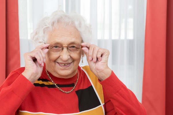 Das Bild zeigt eine weißhaarige Seniorin mit rotem Pullover. Sie trägt eine Brille, die sie links und rechts mit den Händen fasst.