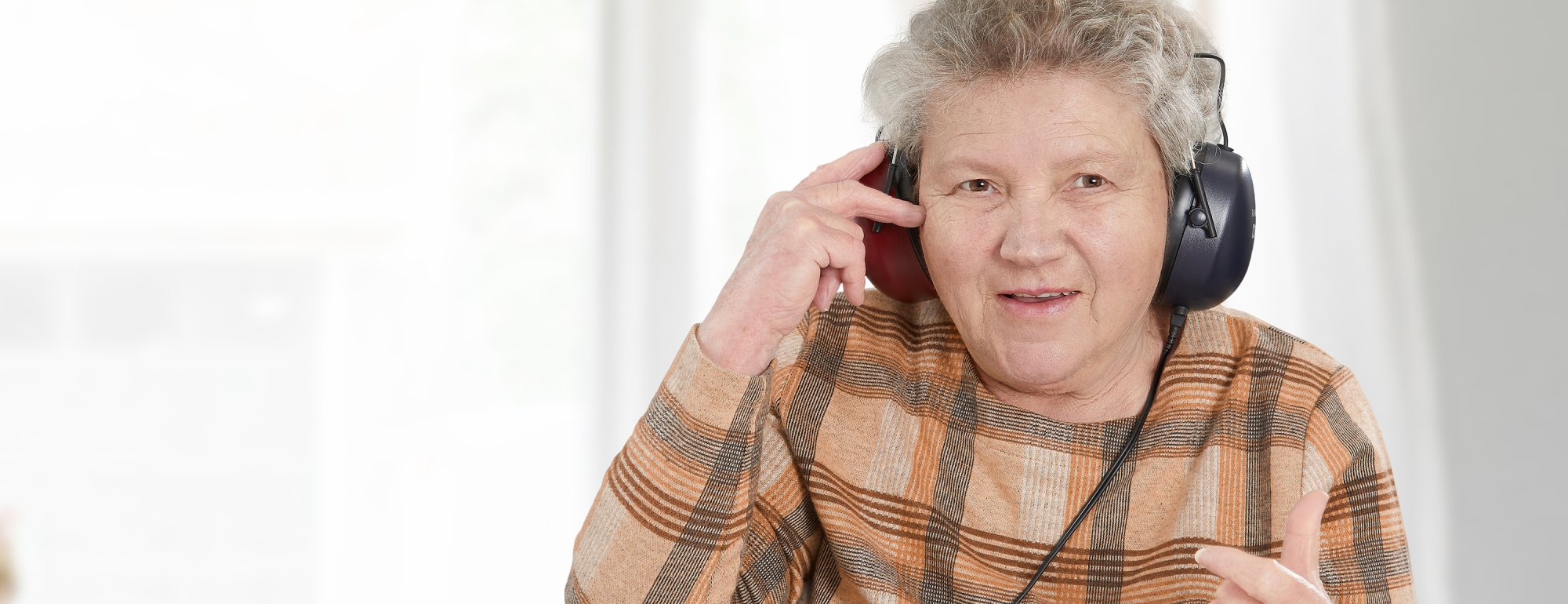 Das Bild zeigt eine Seniorin mit grauen Haaren und einem karierten Oberteil. Sie trägt einen Kopfhörer.