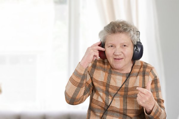 Das Bild zeigt eine Seniorin mit grauen Haaren und einem karierten Oberteil. Sie trägt einen Kopfhörer.