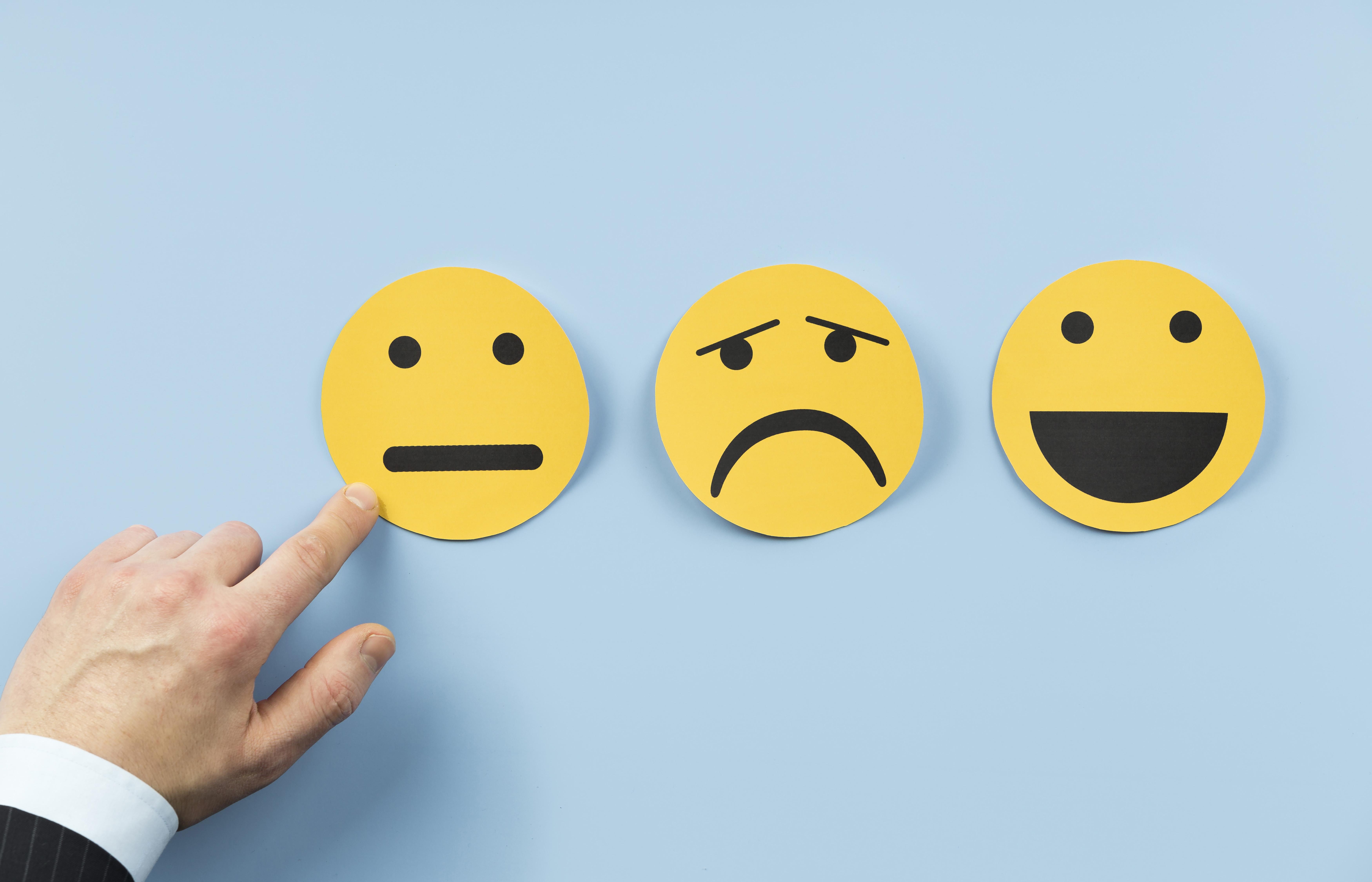 Drei gelbe Smileys auf blauem Hintergrund. Links neutral, in der Mitte traurig, rechts fröhlich. Eine Hand zeigt auf den neutralen Smiley.