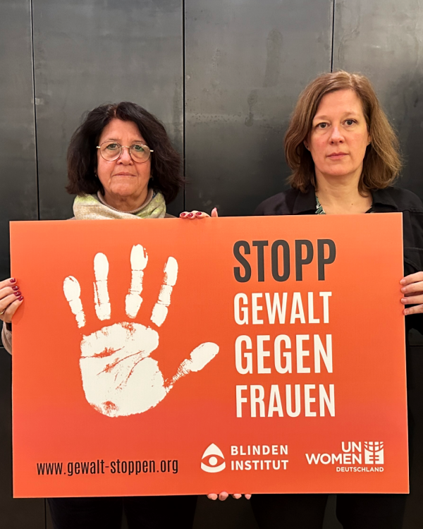 Zwei Frauen halten ein oranges Schild mi einer weißen Handfläche. Daneben steht: Stopp Gewalt gegen Frauen.