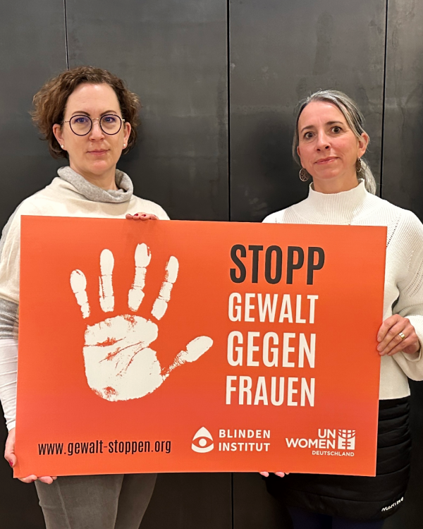 Zwei Frauen halten ein oranges Schild mi einer weißen Handfläche. Daneben steht: Stopp Gewalt gegen Frauen.