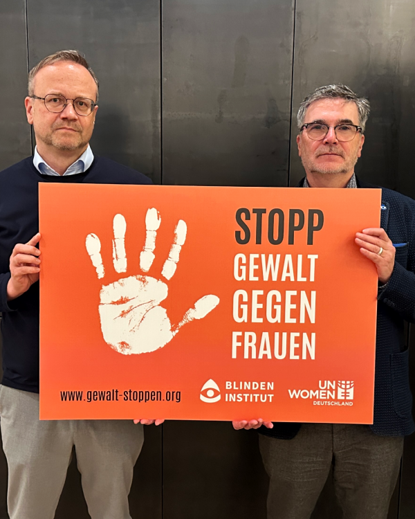Zwei Männer halten ein oranges Schild mi einer weißen Handfläche. Daneben steht: Stopp Gewalt gegen Frauen.