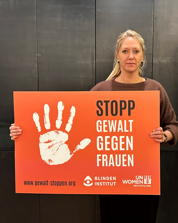 Eine Frau mit blonden Haaren und Zopf hält ein oranges Schild mi einer weißen Handfläche. Daneben steht: Stopp Gewalt gegen Frauen.