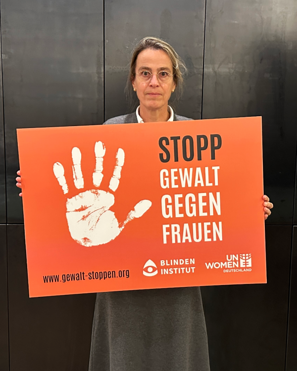 Eine Frau mit Brille hält ein oranges Schild mi einer weißen Handfläche. Daneben steht: Stopp Gewalt gegen Frauen.