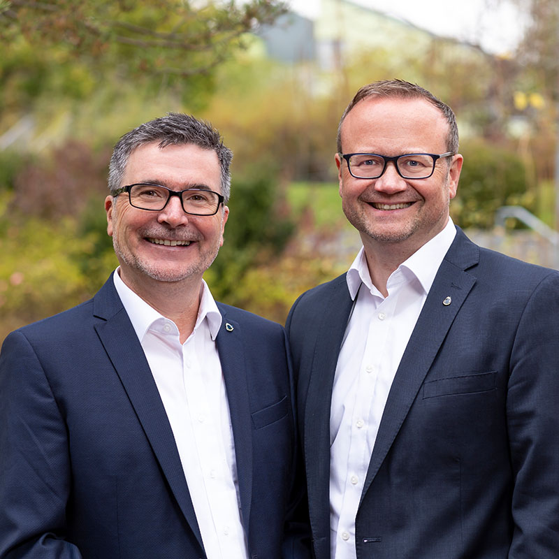 Die Vorstände der Blindeninstitutsstiftung Johannes Spielmann und Dr. Marco Bambach stehen nebeneinander und tragen ein weißes Hemd und ein dunkles Sakko. Im Hintergrund sind Bäume.