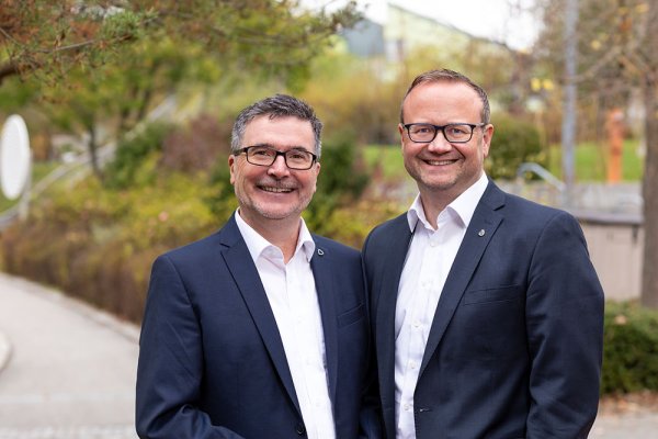 Zwei Männer im Anzug lächeln in die Kamera. Im Hintergrund ist die Außenanlage des Blindeninstituts in Würzburg zu sehen.