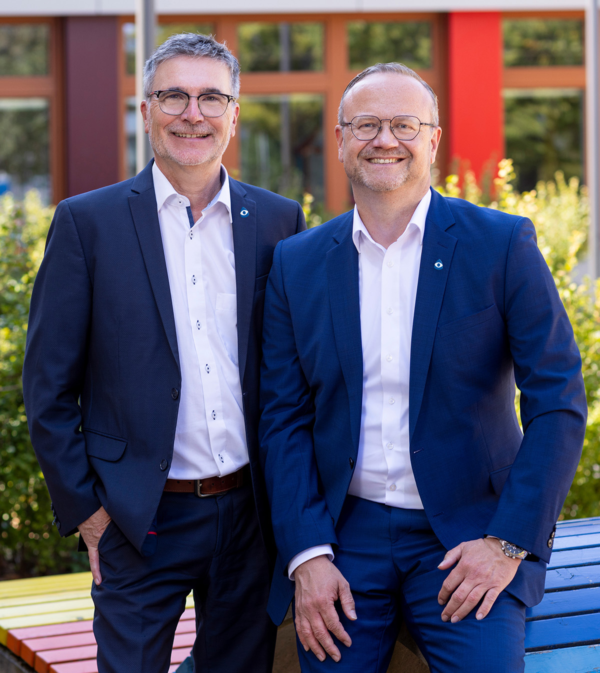 Zwei Männer in blauen Anzügen und weißen Hemden stehen nebeneinander im Freien und lächeln in die Kamera; im Hintergrund grüne Sträucher und eine Fassade mit roten Fensterrahmen, im Vordergrund eine bunt bemalte Holzbank.