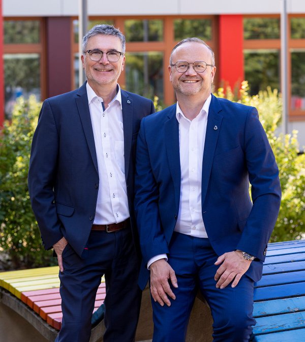 Zwei Männer in blauen Anzügen und weißen Hemden stehen nebeneinander im Freien und lächeln in die Kamera; im Hintergrund grüne Sträucher und eine Fassade mit roten Fensterrahmen, im Vordergrund eine bunt bemalte Holzbank.