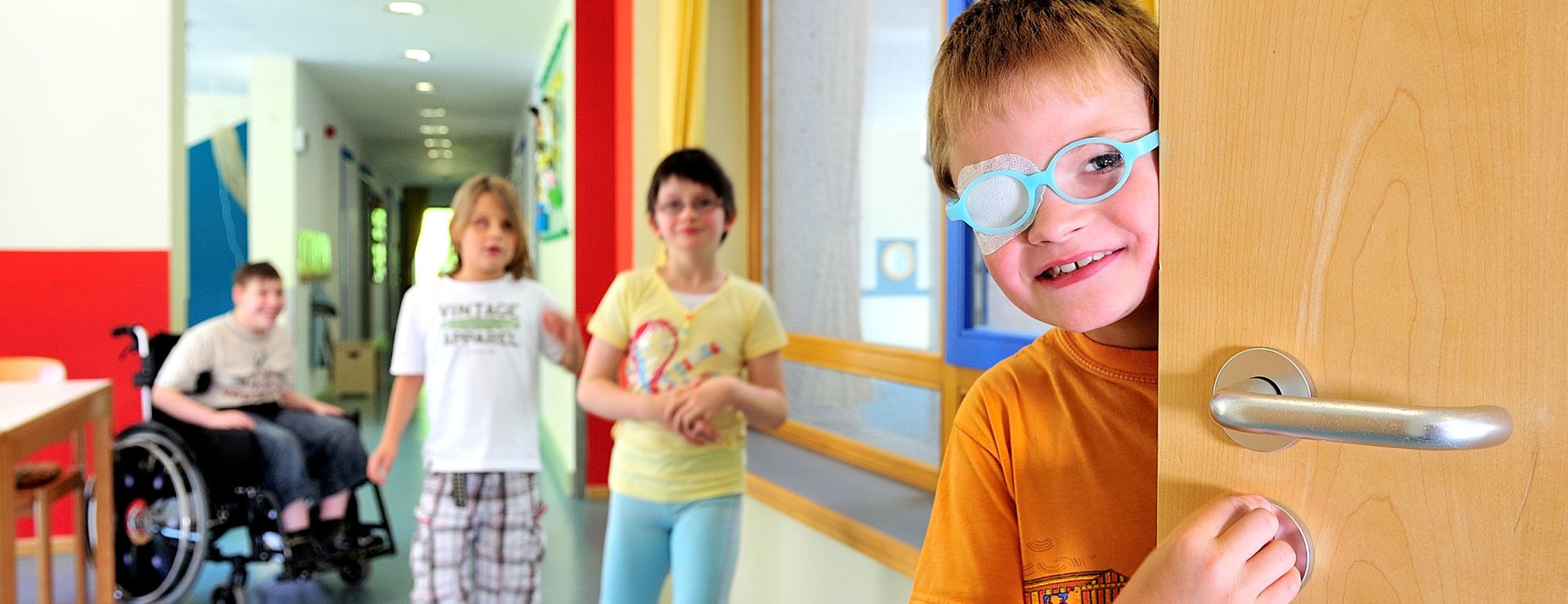 Ein Junge mit Augenpflaster und Brille steht hinter einer Tür und lächelt in die Kamera. Hinter ihm sind drei weitere Kinder, eines von ihnen sitzt im Rollstuhl.