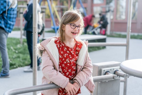 Ein Mädchen mit Brille und Zahnlücke steht an einem Metallgeländer im Freien und lacht. Im Hintergrund sind unscharf weitere Menschen und Spielgeräte zu sehen.