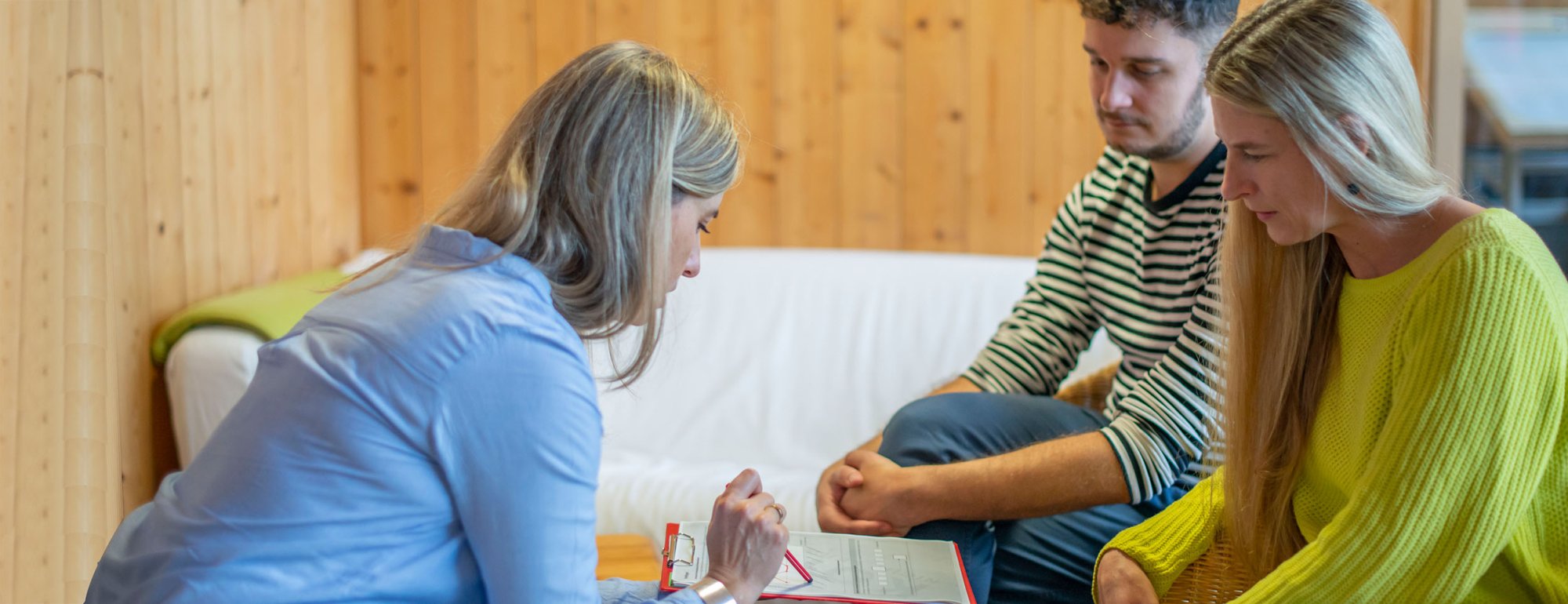 Links sitzt eine Frau vornübergebeut auf einem Stuhl. Sie hält ein Clipboard mit einem Formular in der einen Hand. In der anderen Hand hält sie einen Stiftung, mit dem Sie auf das Formular zeigt. Ihr gegenüber sitzen eine junge Frau und ein junger Mann, die auf das Formular blicken.